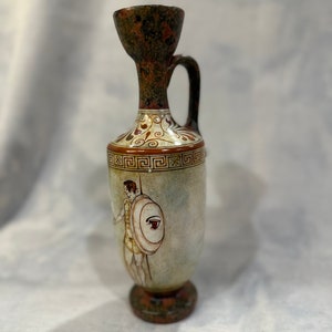 May include: A small, brown and white ceramic vase with a handle and a painted design of a warrior with a shield. The vase has a geometric pattern around the top.