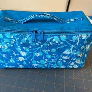 May include: A blue zippered pouch with a white floral pattern and a blue handle. The pouch is sitting on a blue and white gridded surface.