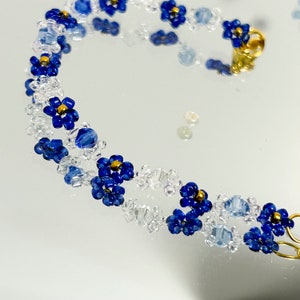 May include: A delicate bracelet made with blue and clear glass beads, featuring a repeating flower design. The bracelet has a gold clasp.