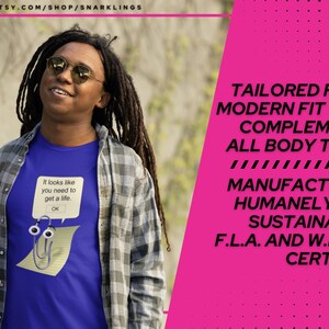 May include: A blue t-shirt with a white paperclip graphic and the text "It looks like you need to get a life. OK." The shirt is being worn by a person with long dreadlocks and a plaid shirt. The background is a blurred image of trees and foliage. The text "TAILORED FOR A MODERN FIT THAT COMPLEMENTS ALL BODY TYPES MANUFACTURED HUMANELY AND SUSTAINABLY. F.L.A. AND W.R.A.P. CERTIFIED" is in white text on a pink background.