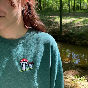 May include: Teal green sweatshirt with an embroidered mushroom design. The design features two mushrooms with red caps and white stems, set on a patch of green grass. The sweatshirt is made of a soft, comfortable material.