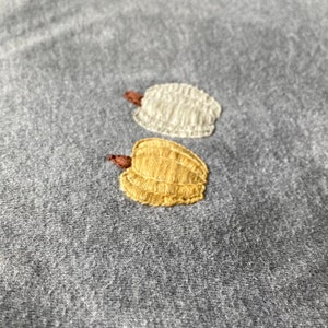 May include: Two embroidered pumpkins on a gray fabric background. One pumpkin is yellow and the other is white.