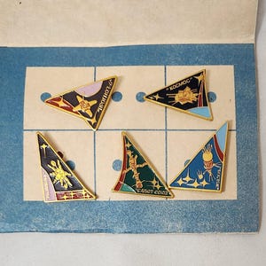 May include: A collection of five vintage triangular enamel pins with gold trim, featuring space-themed designs and Cyrillic text. The pins are arranged on a blue and cream-colored background, likely from a collectible set.
