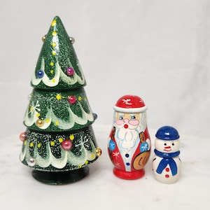 May include: A trio of hand-painted wooden Christmas decorations. A green tiered Christmas tree with colorful ornaments, a Santa Claus figure in red and white, and a small snowman with a blue hat and scarf.