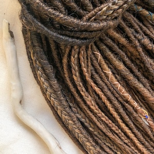 May include: A bundle of brown and blonde synthetic dreadlocks. The dreadlocks are arranged in a loose pile and have a natural, textured appearance.