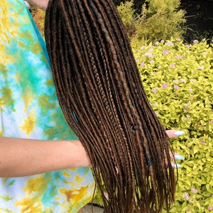 May include: A long, brown synthetic dreadlock wig with blue and copper accents. The wig is held in the hand of a person wearing a tie-dye shirt.