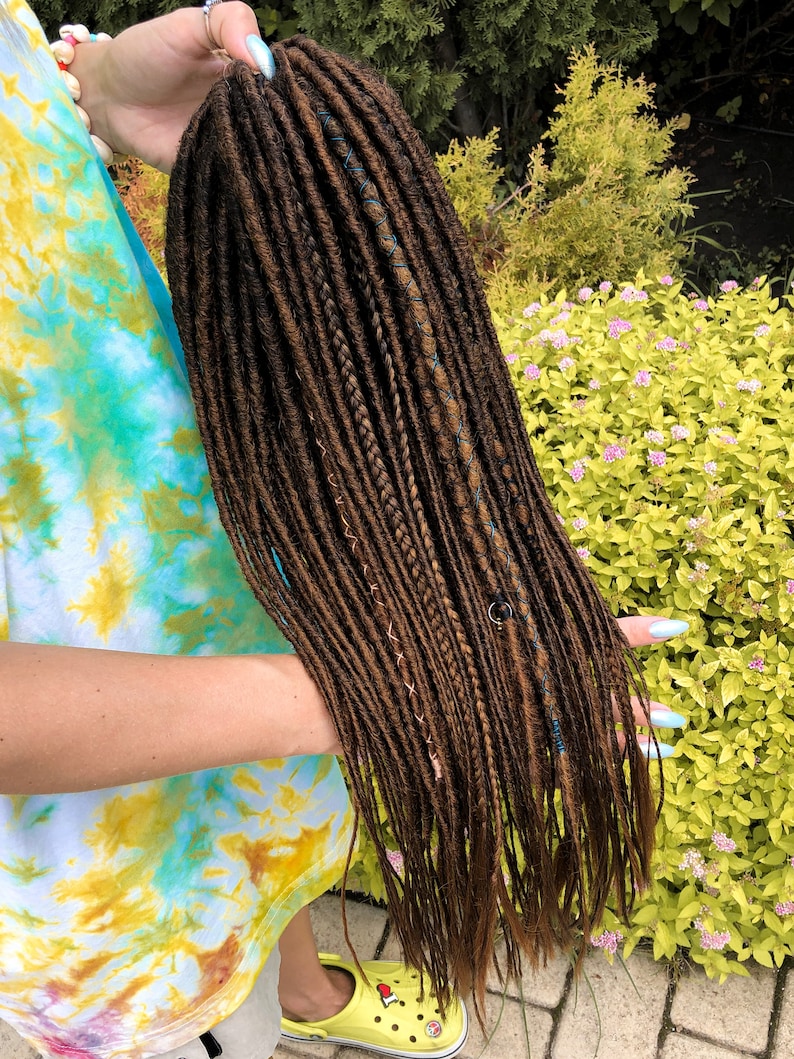 May include: A long, brown synthetic dreadlock wig with blue and copper accents. The wig is held in the hand of a person wearing a tie-dye shirt.