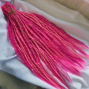Puede incluir: Un conjunto de rastas de color rosa vibrante con un efecto degradado, que pasa de un rosa más claro a un tono más brillante en los extremos. Las rastas están atadas con hilo.