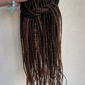 May include: A long brown synthetic dreadlock wig with brown and blue accents. The wig is tied up in a bun at the top.