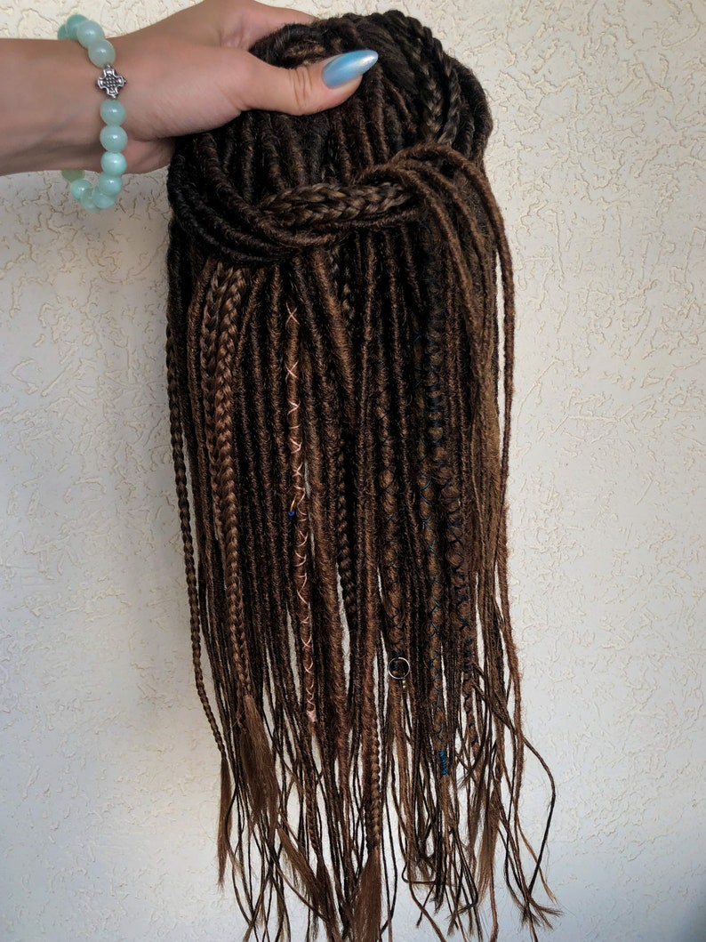 May include: A long brown synthetic dreadlock wig with brown and blue accents. The wig is tied up in a bun at the top.