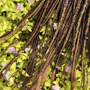May include: A set of brown synthetic dreadlocks with blue and white accents. The dreadlocks are arranged on a bed of green leaves and pink flowers.
