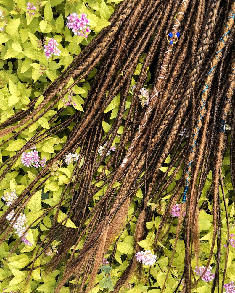 May include: A set of brown synthetic dreadlocks with blue and white accents. The dreadlocks are arranged on a bed of green leaves and pink flowers.