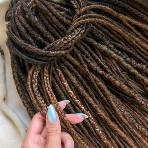 May include: A close-up of a hand holding a bundle of brown and blonde synthetic dreadlocks. The dreadlocks are tied together at the top and have a few brown and blue stitching details.
