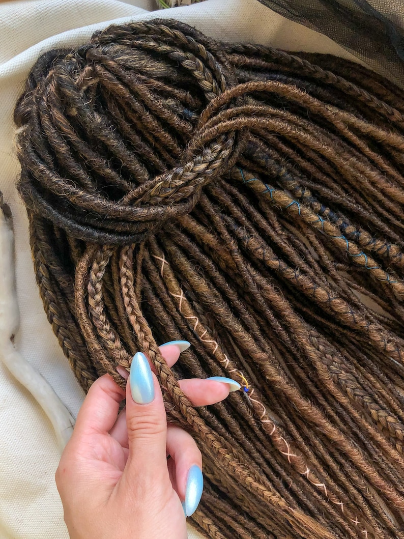 May include: A close-up of a hand holding a bundle of brown and blonde synthetic dreadlocks. The dreadlocks are tied together at the top and have a few brown and blue stitching details.