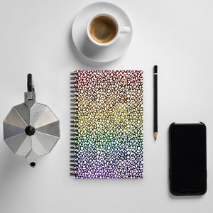 May include: A white notebook with a rainbow speckled pattern, a black pencil, a silver coffee pot, a white coffee cup with coffee, and a black smartphone.