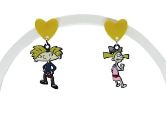 May include: A pair of dangle earrings featuring yellow heart-shaped studs. One earring displays a cartoon boy with blonde hair in a blue shirt, while the other shows a cartoon girl with blonde hair in a pink dress and purple bow.