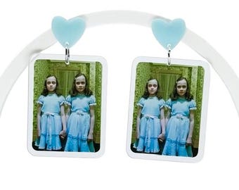 May include: A pair of dangle earrings featuring light blue heart-shaped studs attached to rectangular charms. Each charm displays a photographic print of two identical children wearing matching light blue dresses in a room with yellow wallpaper.