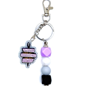 May include: A silver keychain with a charm that reads "THE WAND CHOOSES THE WIZARD" in purple and white. The keychain has a beaded accent in shades of purple, white, gray, and black, and a silver-colored clasp.