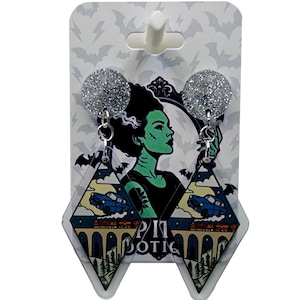 May include: A pair of dangle earrings featuring a Bride of Frankenstein design. The earrings have silver glitter studs and diamond-shaped pendants with a vintage scene and the text "PIT DOTIC". The background is a card with bat silhouettes.