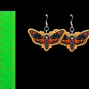 May include: A pair of moth-shaped drop earrings featuring a skull pattern on the thorax, coloured in orange, black, and blue. The earrings are displayed next to a green ruler showing a width of approximately 7.6 cm for each moth charm.