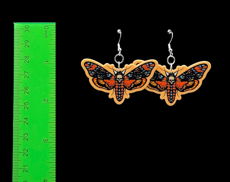 May include: A pair of moth-shaped drop earrings featuring a skull pattern on the thorax, coloured in orange, black, and blue. The earrings are displayed next to a green ruler showing a width of approximately 7.6 cm for each moth charm.