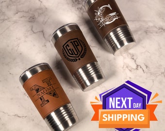 May include: Three stainless steel tumblers with brown leather wraps. Each tumbler has a different personalized design. The designs include a floral monogram, a monogram with a laurel wreath, and a monogram with a decorative design. The tumblers are on a marble surface.  Next Day Shipping is written on a purple and orange graphic.
