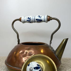 May include: A copper kettle with a brass spout and handle. The handle is white ceramic with blue floral designs. The lid is brass with a white and blue ceramic knob. The kettle is sitting on a beige surface.