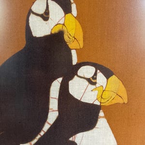 May include: A textile wall hanging featuring a graphic illustration of two puffins against a solid burnt orange background. The birds are depicted with black plumage, white facial patches, and prominent yellow beaks, accented by thin red lines.