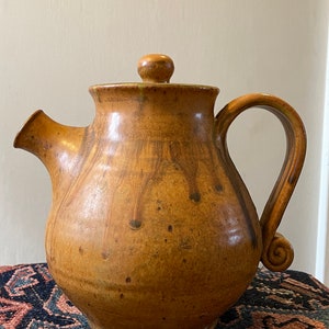 May include: A brown ceramic teapot with a rounded body, a lid with a knob, and a curved handle. The teapot has a speckled glaze and is sitting on a colorful rug.