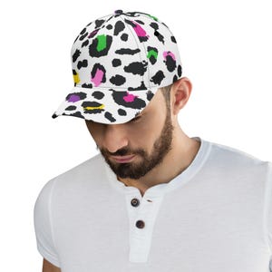Pode incluir: Um bon&eacute; de beisebol branco com um padr&atilde;o de estampa de leopardo preto e branco. A estampa apresenta respingos de rosa, verde e amarelo.