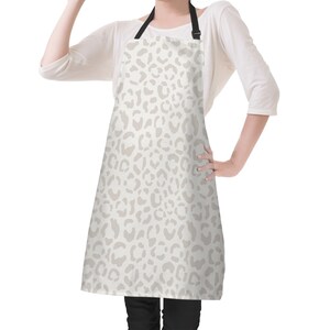 May include: A beige apron featuring a light brown leopard print design. The apron has a black adjustable neck strap and a black waist tie. The apron is worn by a person wearing a white long-sleeved shirt and black trousers.
