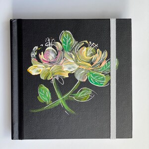 Könnte beinhalten: Ein schwarzes Journal mit einem weißen Gummibandverschluss und einem handgemalten Blumendesign mit gelben, grünen und rosa Blüten.