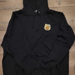 May include: Black hooded sweatshirt with a drawstring and a small patch featuring a cartoon dog on fire. The sweatshirt has a kangaroo pocket and a Russell Athletic logo on the lower left.