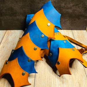 Könnte beinhalten: Handgefertigte Lederpanzerung in Blau- und Orangetönen, mit Metallnieten befestigt. Das segmentierte Design deutet auf ein Schulter- oder Brustteil mit einer rustikalen, mittelalterlichen Ästhetik hin. Das Leder hat eine natürliche Maserung und Textur.
