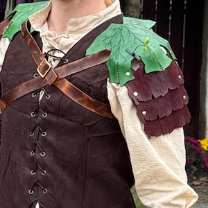May include: A brown leather vest with a crisscross lace-up front and a brown leather strap across the chest. The shoulder armor is green and burgundy, with a leaf design. The sleeves are cream-coloured.