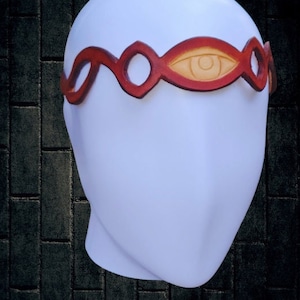 May include: A red leather headband with a decorative eye design. The eye is carved into the leather and has a yellow center.