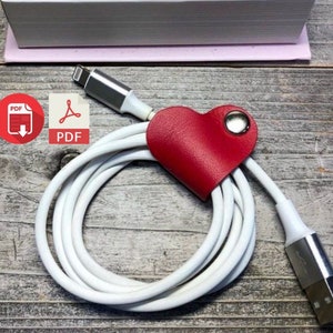 May include: A white charging cable with a red heart-shaped leather cord organizer. The cable is wrapped around the organizer and the end of the cable is plugged into a device. The image includes the text "PDF" in a red circle with a white download arrow.