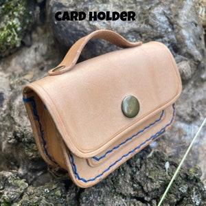 May include: A light brown leather card holder with a flap closure and a blue stitched border. The holder has a brass button closure and a leather carrying handle.