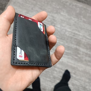 May include: A black leather card holder with a stitched edge. The holder has two slots for cards, one of which is visible and shows a red card with the word "Visa" printed on it.