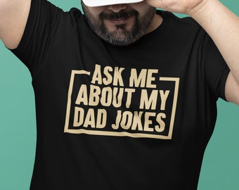 Könnte beinhalten: Schwarzes T-Shirt mit dem Text "ASK ME ABOUT MY DAD JOKES" in beiger Schrift mit Umrandung. Der Text ist mittig auf dem Shirt platziert. Die Person trägt eine weiße Baseballkappe.