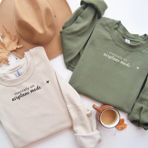 K&ouml;nnte beinhalten: Zwei Sweatshirts mit Rundhalsausschnitt, eines in Beige und eines in Olivgr&uuml;n, mit dem Aufdruck "mentally on airplane mode" auf der Vorderseite. Ein brauner Hut und eine Tasse Kaffee sind ebenfalls im Bild. Die Sweatshirts haben ein Herz- und Flugzeugmotiv.