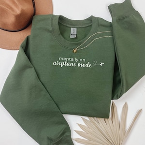 K&ouml;nnte beinhalten: Ein olivgr&uuml;nes Sweatshirt mit dem wei&szlig;en Text "mentally on airplane mode". Eine goldene Halskette mit Anh&auml;nger liegt &uuml;ber dem Sweatshirt. Ein brauner Hut ist in der oberen linken Ecke zu sehen.
