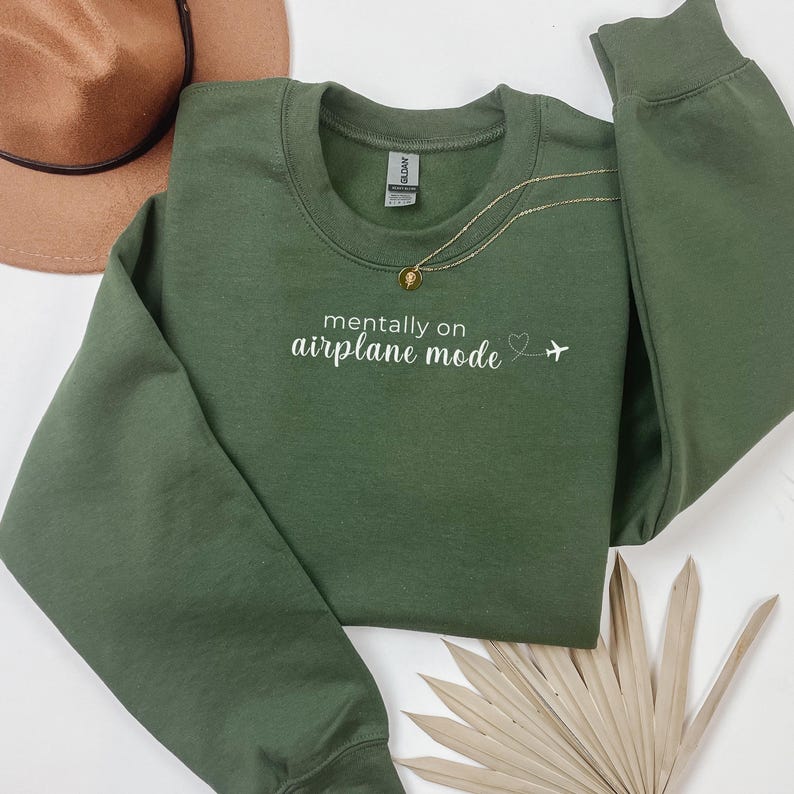 K&ouml;nnte beinhalten: Ein olivgr&uuml;nes Sweatshirt mit dem wei&szlig;en Text "mentally on airplane mode". Eine goldene Halskette mit Anh&auml;nger liegt &uuml;ber dem Sweatshirt. Ein brauner Hut ist in der oberen linken Ecke zu sehen.