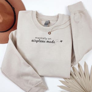 K&ouml;nnte beinhalten: Beiges Sweatshirt mit Rundhalsausschnitt und dem schwarzen Text "mentally on airplane mode". Ein kleines Herz und ein Flugzeug-Symbol befinden sich neben dem Text. Eine goldene Halskette mit Anh&auml;nger liegt auf dem Sweatshirt.