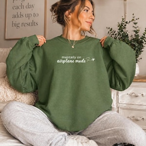 K&ouml;nnte beinhalten: Ein olivgr&uuml;nes Sweatshirt mit dem Text "mentally on airplane mode" und einer kleinen Flugzeuggrafik. Das Sweatshirt wird mit grauen Jogginghosen kombiniert. Im Hintergrund ist ein gerahmter Druck mit dem Text "A little progress each day adds up to big results" zu sehen.