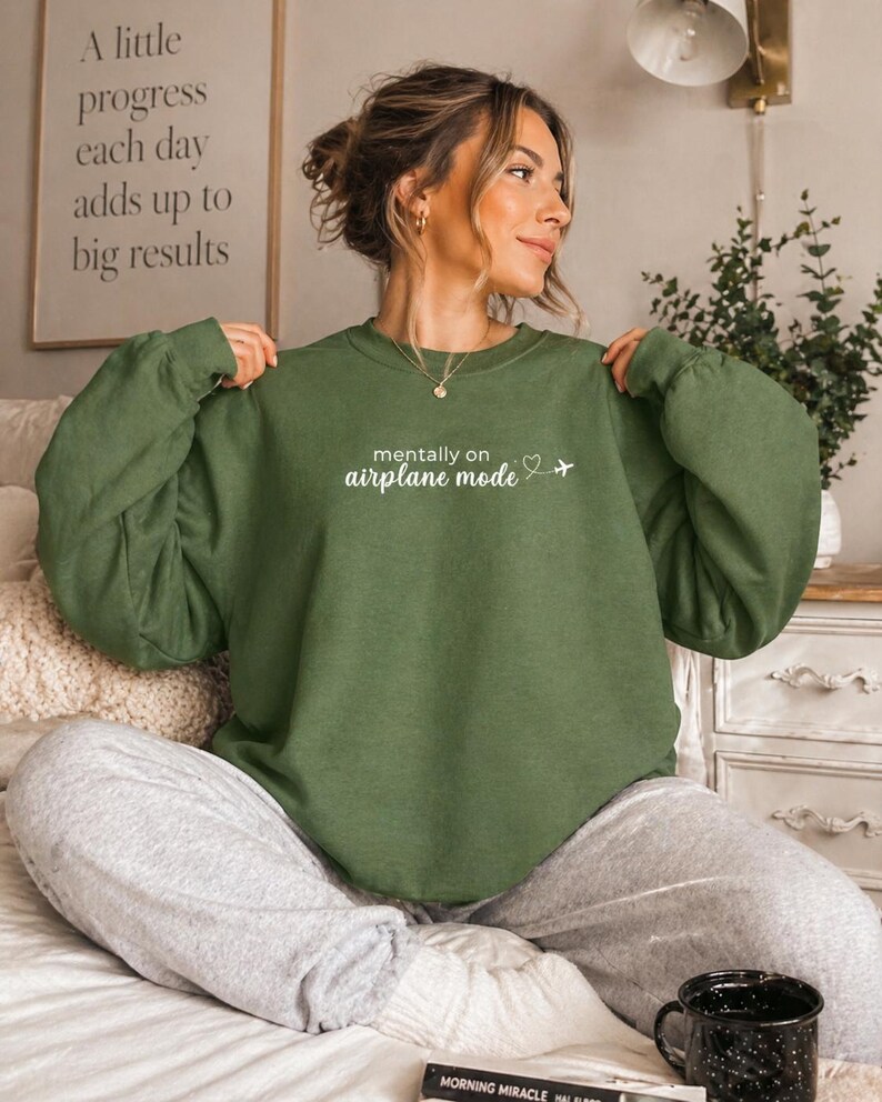 K&ouml;nnte beinhalten: Ein olivgr&uuml;nes Sweatshirt mit dem Text "mentally on airplane mode" und einer kleinen Flugzeuggrafik. Das Sweatshirt wird mit grauen Jogginghosen kombiniert. Im Hintergrund ist ein gerahmter Druck mit dem Text "A little progress each day adds up to big results" zu sehen.
