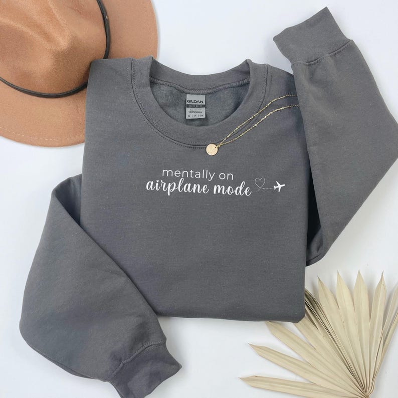 K&ouml;nnte beinhalten: Ein graues Sweatshirt mit dem wei&szlig;en Schriftzug "mentally on airplane mode". Eine goldene Halskette mit einem kleinen Anh&auml;nger liegt auf dem Sweatshirt. Ein brauner Hut und eine getrocknete Pflanze sind im Hintergrund.