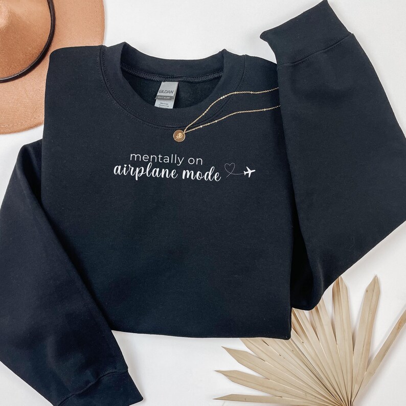 K&ouml;nnte beinhalten: Schwarzes Sweatshirt mit Rundhalsausschnitt und dem wei&szlig;en Schriftzug "mentally on airplane mode". Ein kleines Herz und ein Flugzeug-Symbol befinden sich neben dem Text. Eine goldene Halskette liegt auf dem Sweatshirt. Ein beiger Hut ist im Hintergrund.