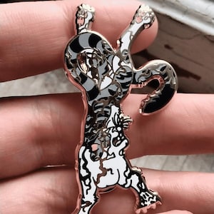 May include: A detailed enamel pin featuring a stylized, abstract figure with a black, white, and gray pattern. The pin has a rose gold outline and is held in a hand. The design is intricate and artistic.
