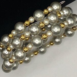 May include: A multi-strand bracelet featuring large, silvery-gray faux pearls interspersed with smaller, gold-toned beads. The bracelet is strung on elastic and has a simple clasp. The design is elegant and suitable for various occasions.